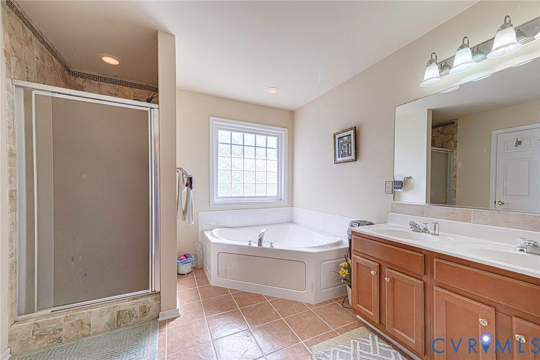 1900 Brilland Court, Glen Allen, VA 23060 Bathroom featuring a corner soaking tub with a block glass window, a walk-in shower with natural stone-look tiling, and a dual vanity with a large mirror and overhead lighting