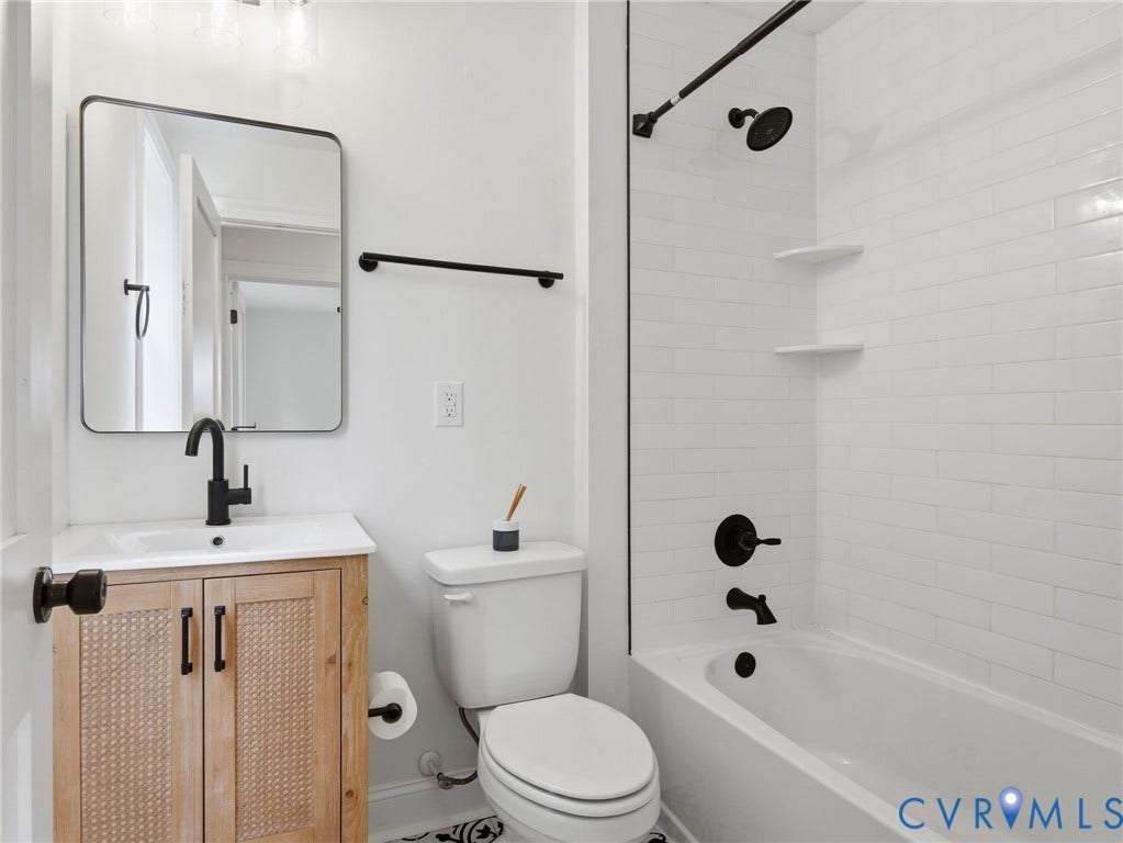 Bathroom featuring a light wood-finish vanity with a rectangular sink, black fixtures, a framed mirror, and a full-size bathtub with white subway tile surround and black shower fixtures