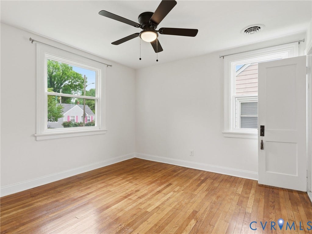 Hardwood flooring throughout this room, featuring two windows with white trim, a white paneled door, and a ceiling fan with light fixture