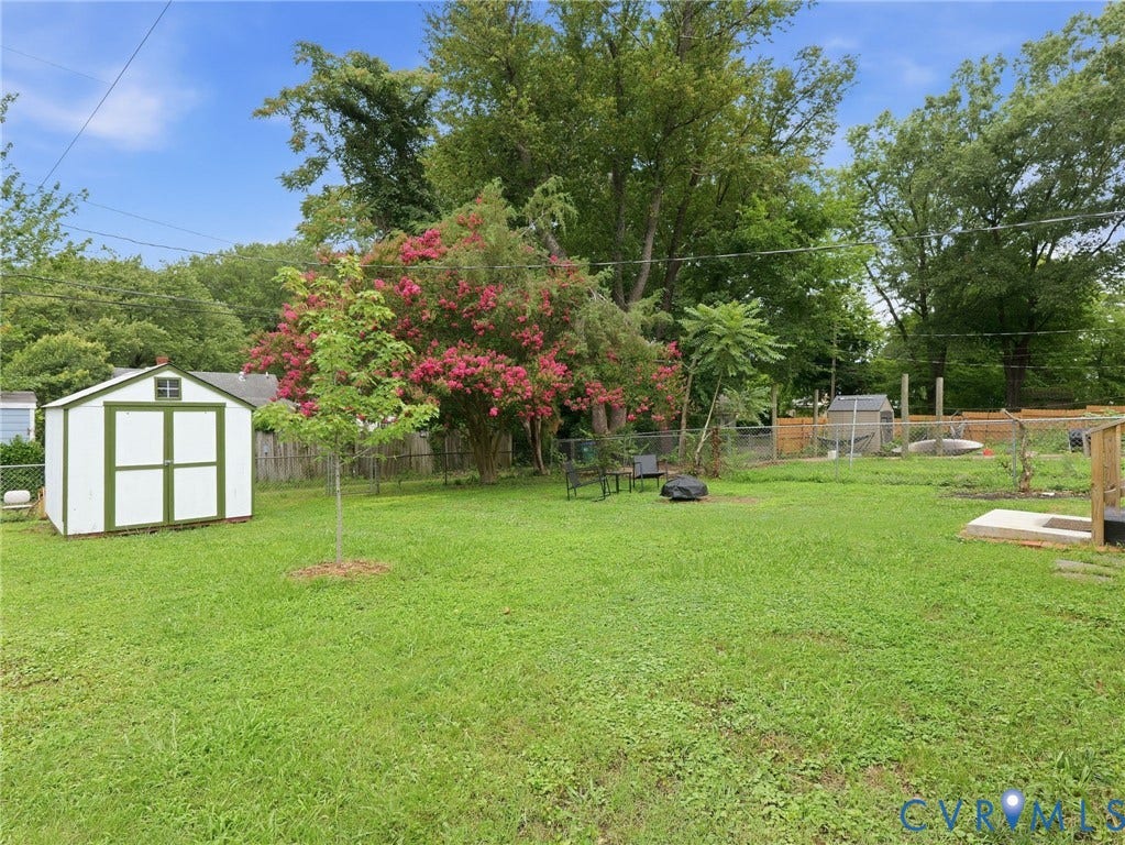 Expansive rear yard featuring a white and green storage shed, mature trees, flowering shrubbery, and a chain-link fence