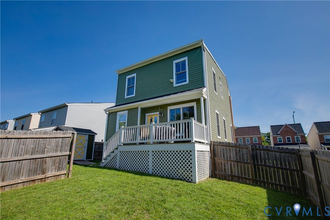 1301 Dance Street, Randolph, Richmond, VA 23220 Rear exterior featuring a two-story design with green siding, an upper-level window, a lower-level deck with white railings and lattice skirting, and a fenced backyard with a green lawn