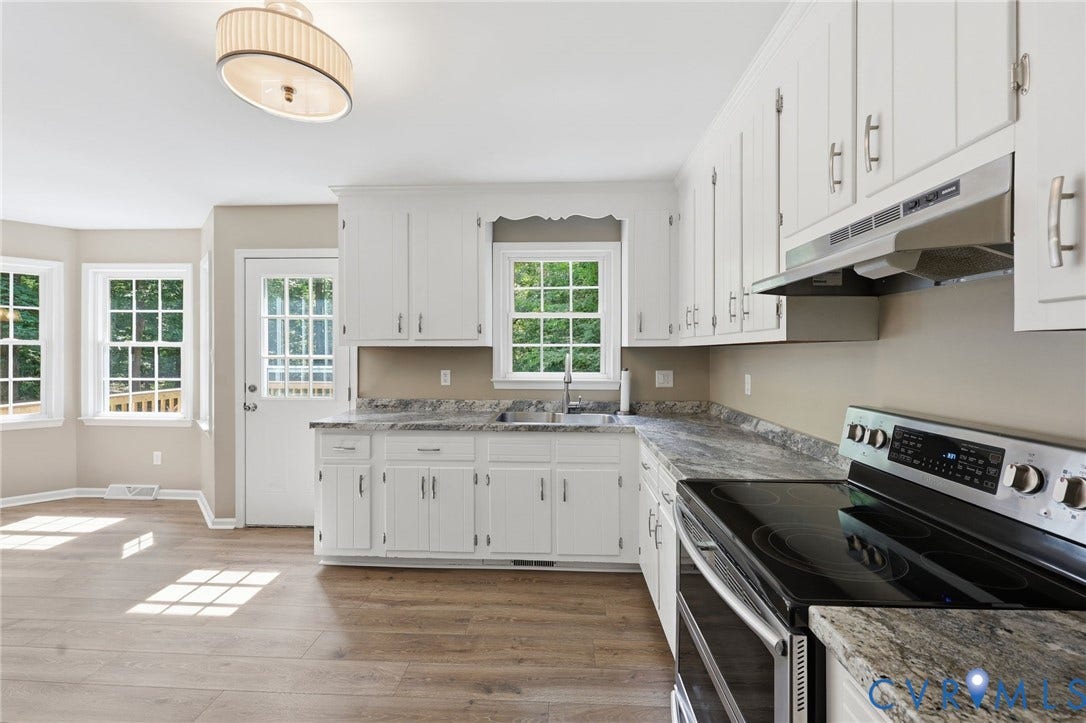 Kitchen featuring white cabinetry, wood-finish flooring, stainless steel range with overhead hood, grey patterned countertops, and multiple windows