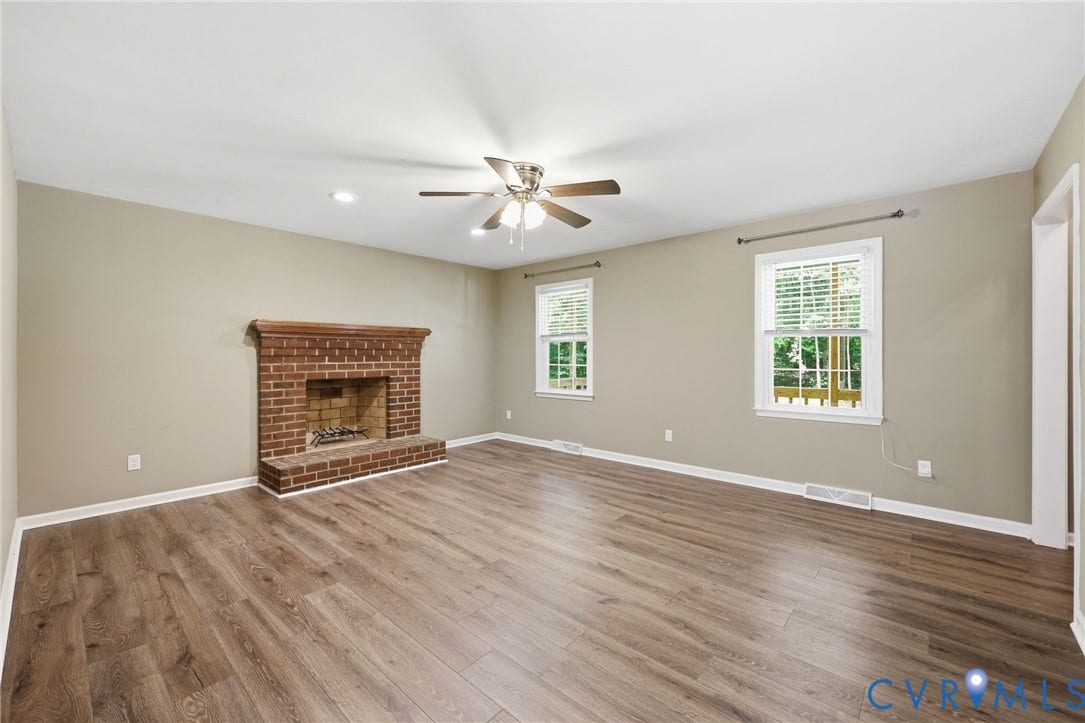 Spacious room featuring wood-finish flooring, a brick fireplace with a wood mantel, and two windows with white blinds