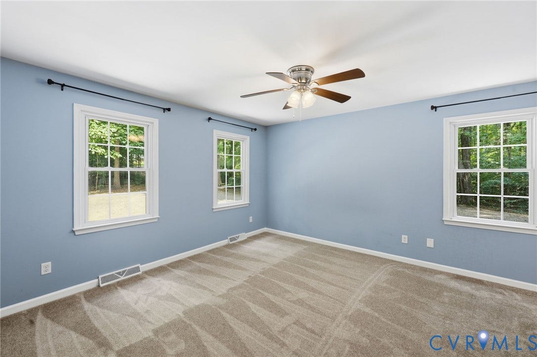 Spacious room featuring multiple large windows, neutral carpet flooring, a ceiling fan with light fixture, and light blue wall finishes