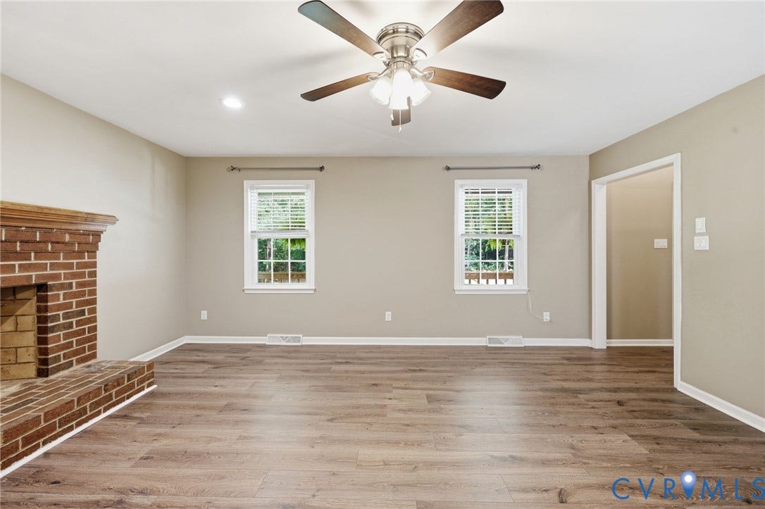 Spacious interior room featuring wood-finish flooring, a brick fireplace with a wood mantle, and a ceiling fan with integrated lighting