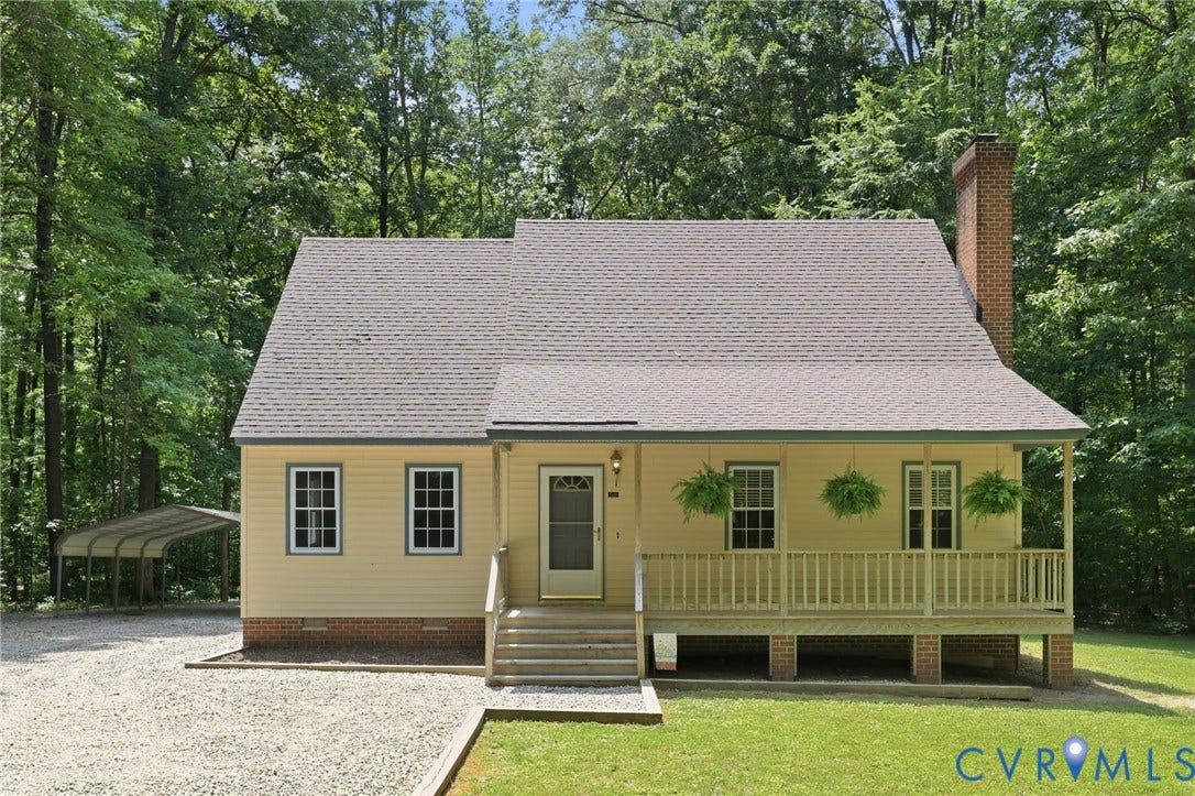 Gable-roof property featuring a light yellow exterior with white trim, a covered front porch with wood railing, a red brick foundation, and a prominent brick chimney