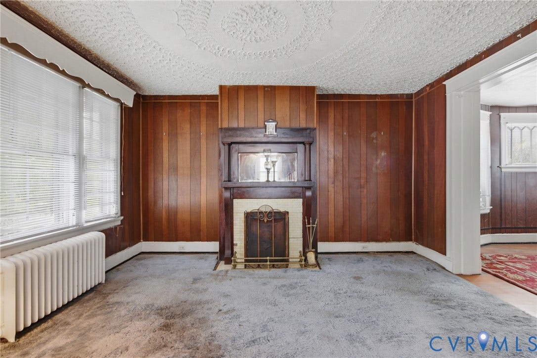 3110 North Avenue, Brookland Park, Richmond, VA 23222 Spacious living room featuring rich wood-paneled walls, original fireplace with decorative mantel and surround, large windows with plantation shutters, radiator heating, and textured ceiling detail