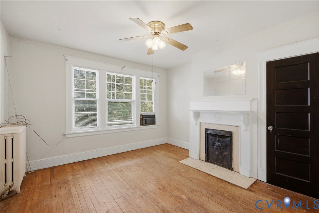 3110 North Avenue, Brookland Park, Richmond, VA 23222 Bright, open living space featuring hardwood flooring, large multi-pane windows with natural light, ceiling fan with integrated lighting, and a fireplace with white surround and dark hearth insert