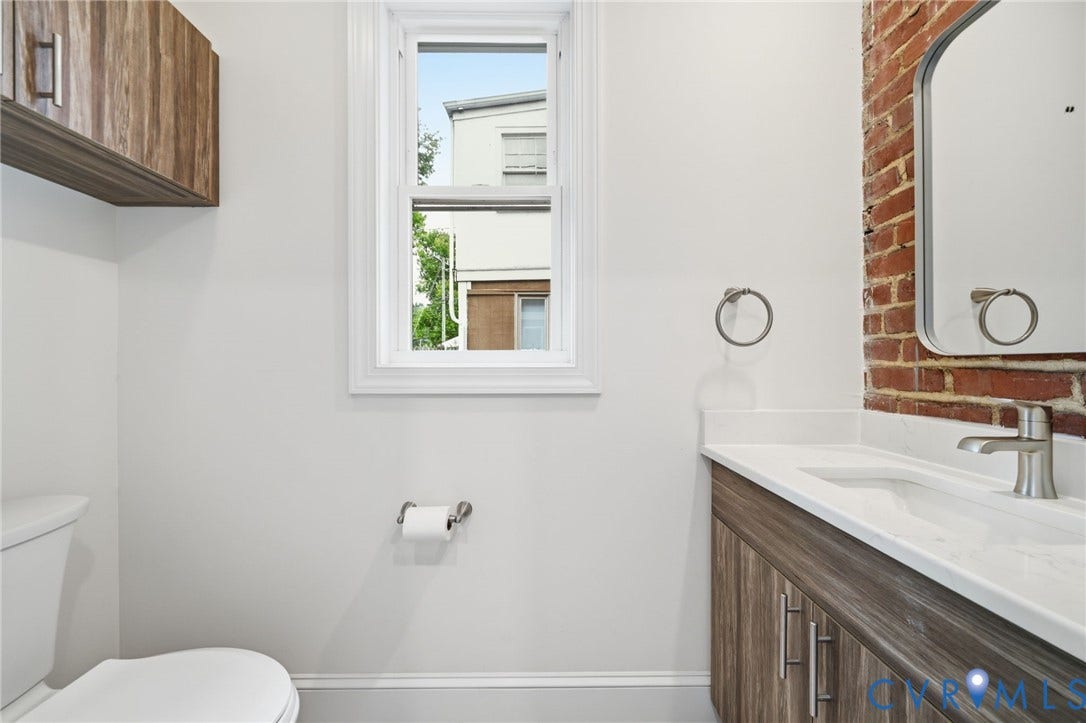 2919 Grayland Avenue, Byrd Park/ Carytown, Richmond, VA 23221 Half Bathroom on first floor, featuring an exposed brick accent wall, a wood-finish vanity with a white countertop and integrated sink, and brushed nickel fixtures