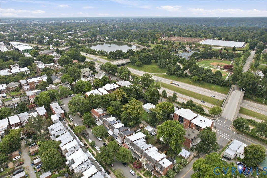 2919 Grayland Avenue, Byrd Park/ Carytown, Richmond, VA 23221 Aerial perspective showcasing a residential area with dense tree cover, a prominent lake, a multi-lane roadway with an overpass, and a sports field