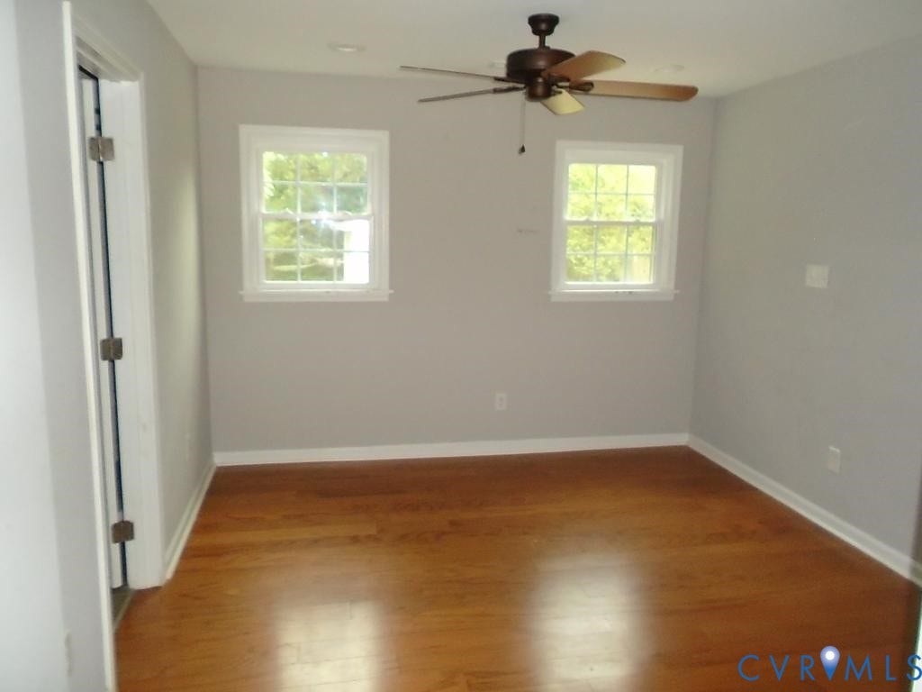 3406 Mike Road, Richmond, VA 23234 Inviting room featuring wood-finish flooring, two large paneled windows with white trim, a dark-toned ceiling fan with light fixture, and light gray wall paint