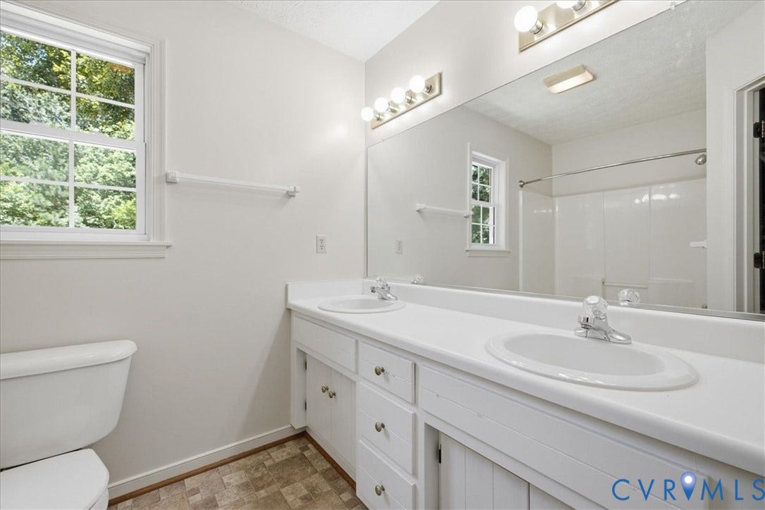 Dual  vanity with integrated sinks, a large wall-mounted mirror, overhead bar lighting, a window with natural light, and patterned flooring