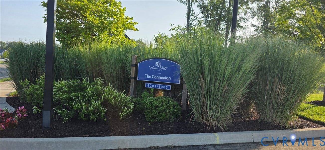 Community entrance sign featuring a blue arched panel and address numbers, surrounded by tall ornamental grasses, varied green plantings, and dark-toned ground cover