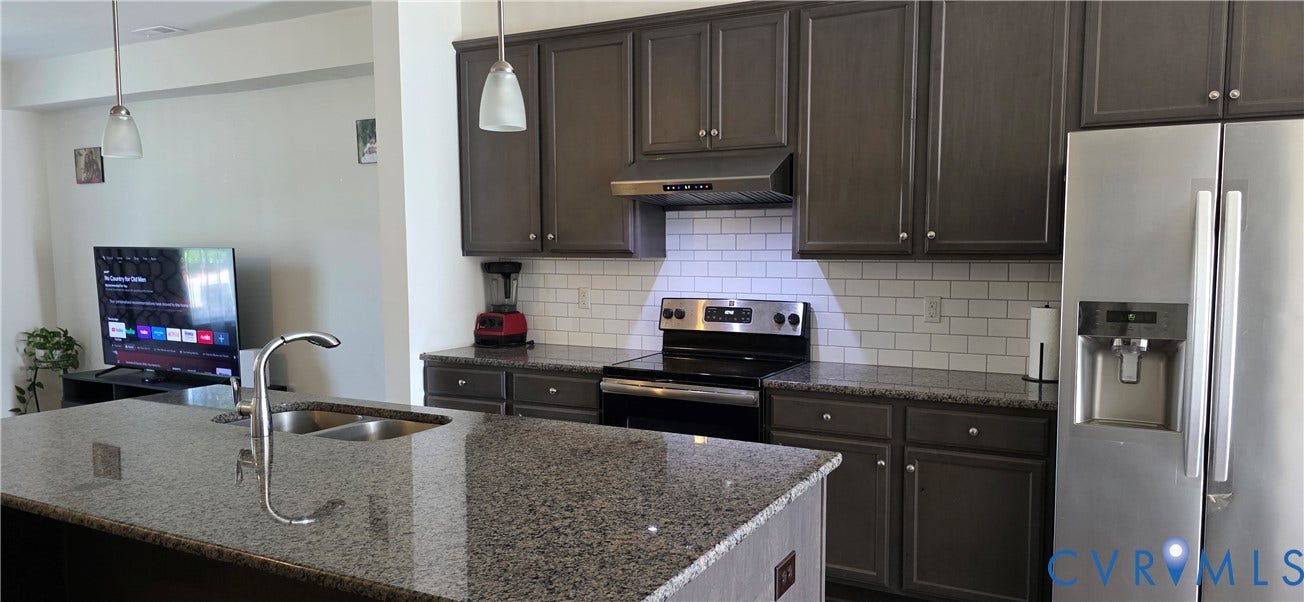 Kitchen featuring an island with dual basin sink and gooseneck faucet, granite countertops, dark wood-finish cabinetry, subway tile backsplash, and stainless steel appliances