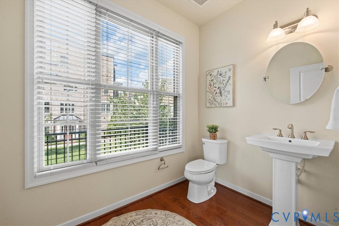 Pedestal sink with brushed nickel faucet, large window with white blinds, round frameless mirror, and overhead vanity lighting