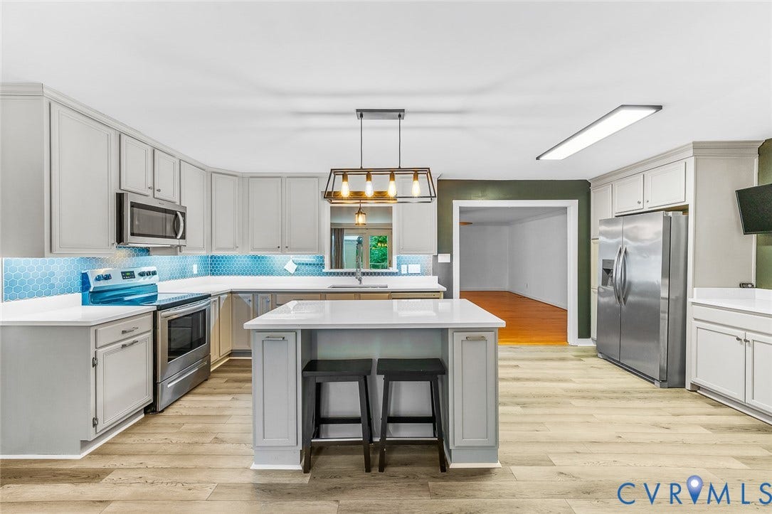 Spacious kitchen featuring an island with seating, stainless steel appliances, light wood-finish flooring, white cabinetry, and a blue hexagonal tile backsplash