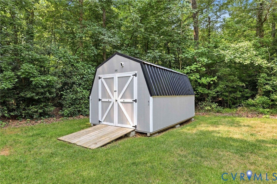 Gray-paneled outbuilding featuring a gambrel roof with black corrugated metal, white double barn doors, and a wood ramp