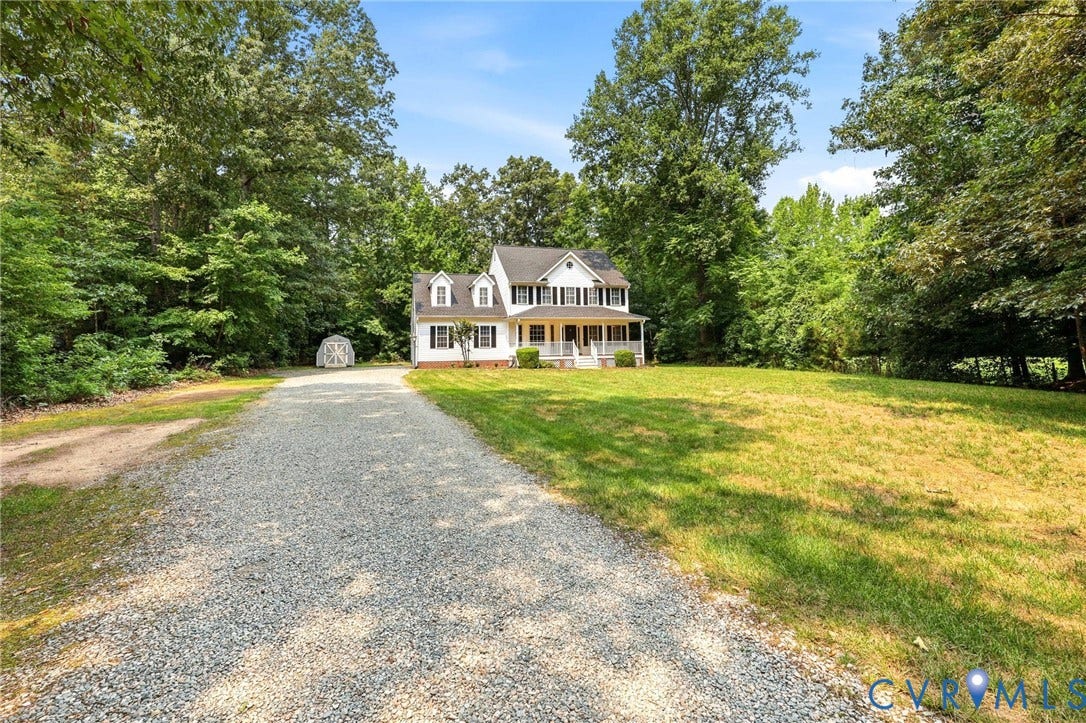 Gravel driveway approaching a white two-story residence with black shutters, a full-width covered porch, and dormer windows