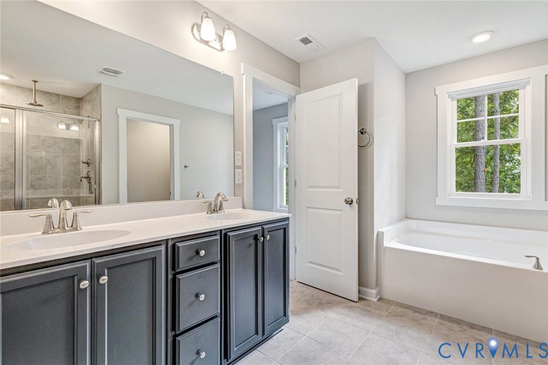 Bathroom featuring a double vanity with dark cabinetry and light countertops, a built-in tub with a window, a glass-enclosed shower with tiled walls, and light-toned floor tiling