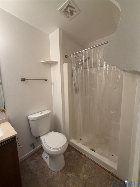 Bathroom featuring a white toilet, enclosed shower with a handheld showerhead, dark wood-finish vanity, light-toned countertop, and patterned floor tiling