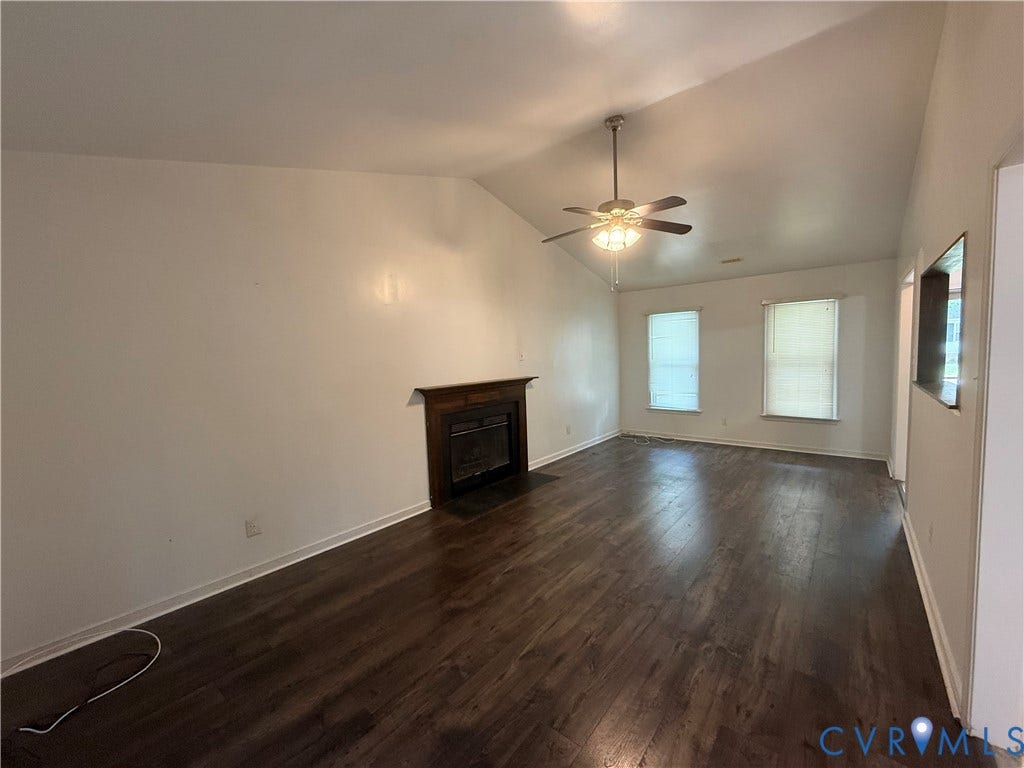 Spacious living area featuring vaulted ceilings, wood-finish flooring, a built-in fireplace with a dark wood mantle, and two windows providing natural illumination
