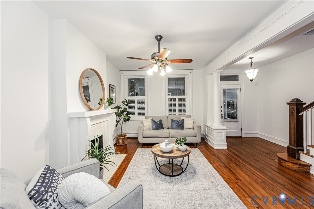 Living area featuring hardwood flooring, a fireplace with a white mantel, ceiling fan, white window shutters, and decorative columns
