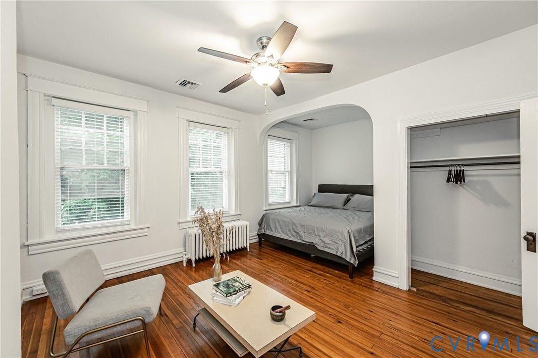 Spacious room featuring rich wood flooring, multiple windows with blinds, a ceiling fan with integrated lighting, and an arched entryway leading to a separate sleeping area