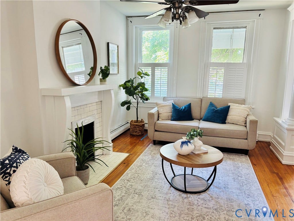 Bright living area featuring a decorative fireplace with white tile surround, wood flooring, and white plantation shutters on double windows