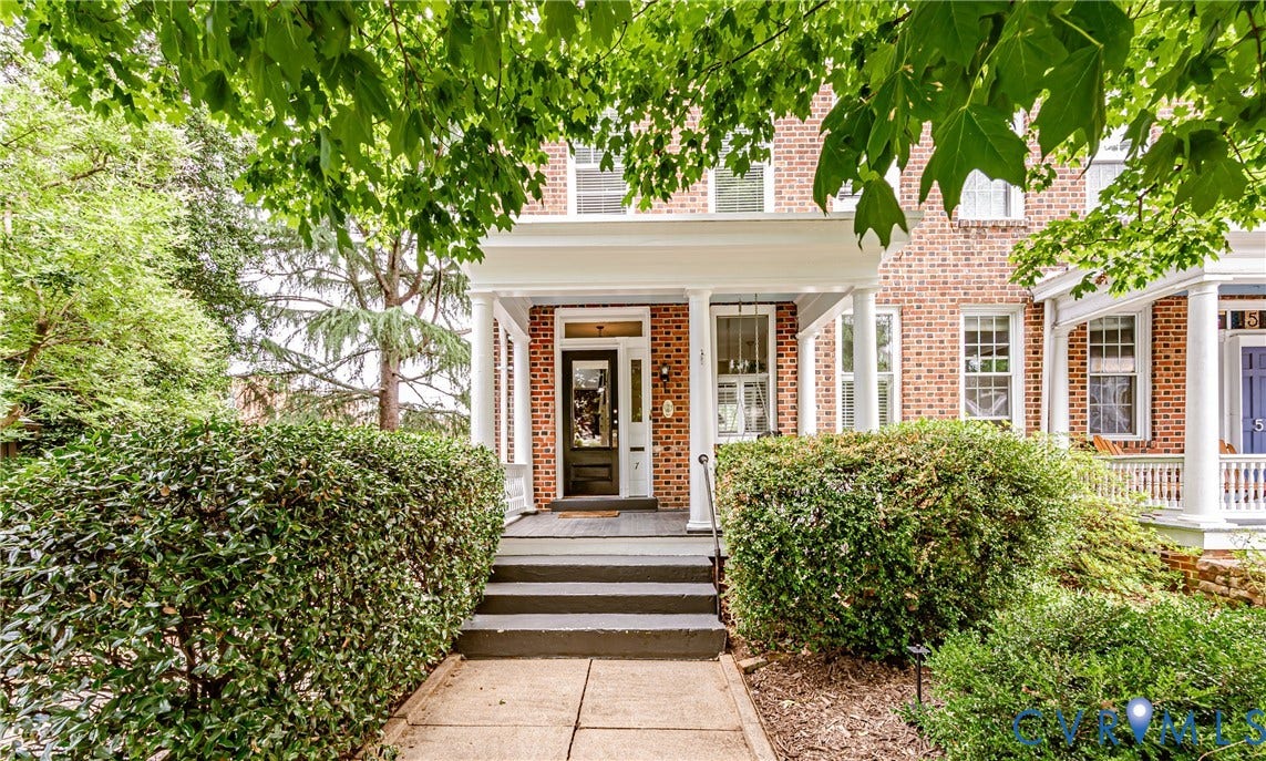 Brick facade residence with a columned front porch, dark paneled entry door with sidelights, and mature front landscaping