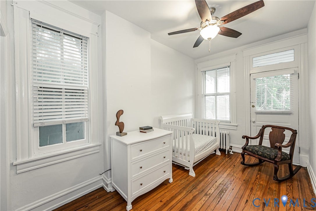 Room with hardwood flooring, white walls, two windows with blinds, a white door with a transom window, and a ceiling fan with light fixture