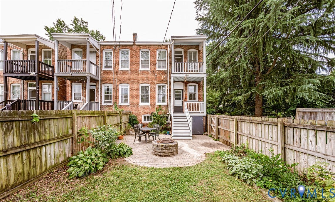 Fenced backyard featuring a paver patio with a built-in fire pit, a mature tree, and a brick exterior with two levels of white-railed balconies