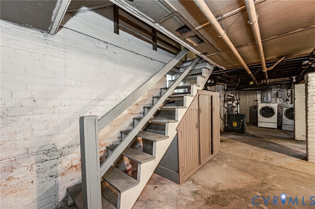 Basement utility area featuring a wooden staircase, exposed overhead piping, concrete flooring, and white-painted brick walls