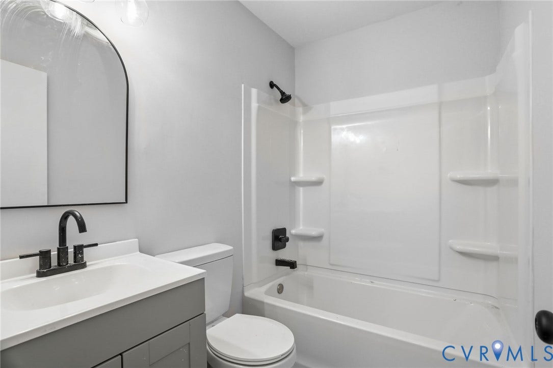 5301 Dalglish Road, Henrico, VA 23223 Bathroom featuring a white bathtub with integrated shelving, matte black fixtures, a contemporary arched mirror, a gray vanity cabinet, and light gray wall paint