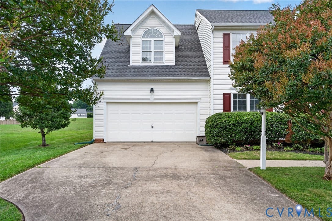 10817 Correnty Drive, Glen Allen, VA 23059 White siding exterior with a two-car garage, concrete driveway, dormer window with an arched transom, dark shutters, and mature landscaping