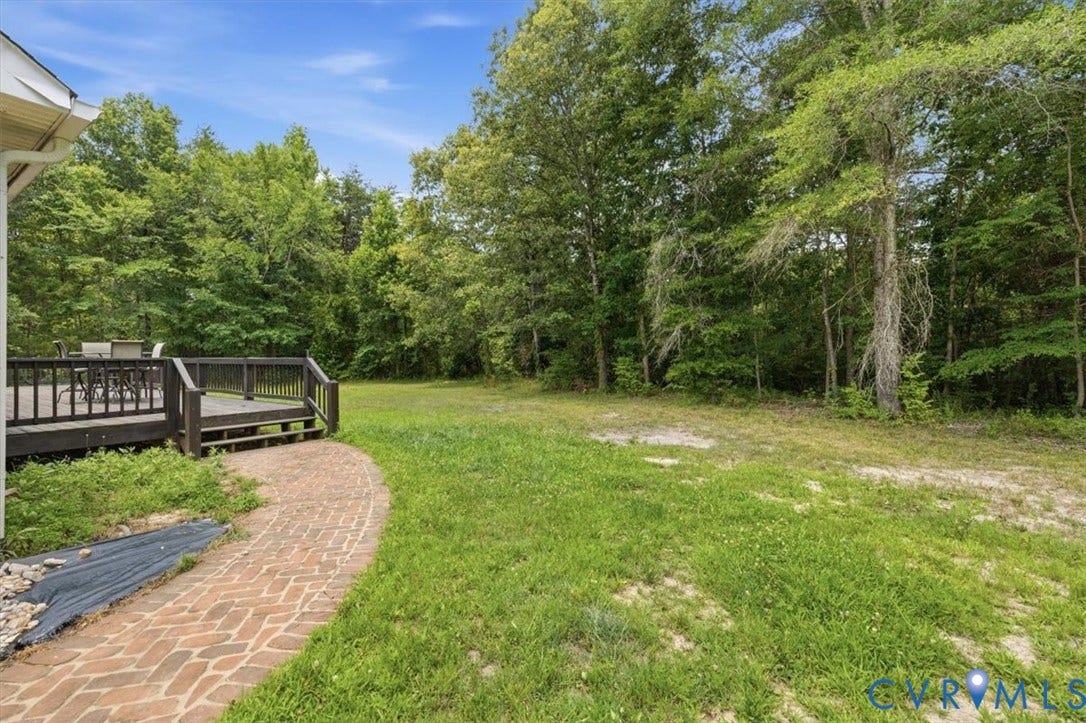 Expansive green space featuring a wooden deck with railing, a brick paver pathway, and mature tree coverage along the perimeter