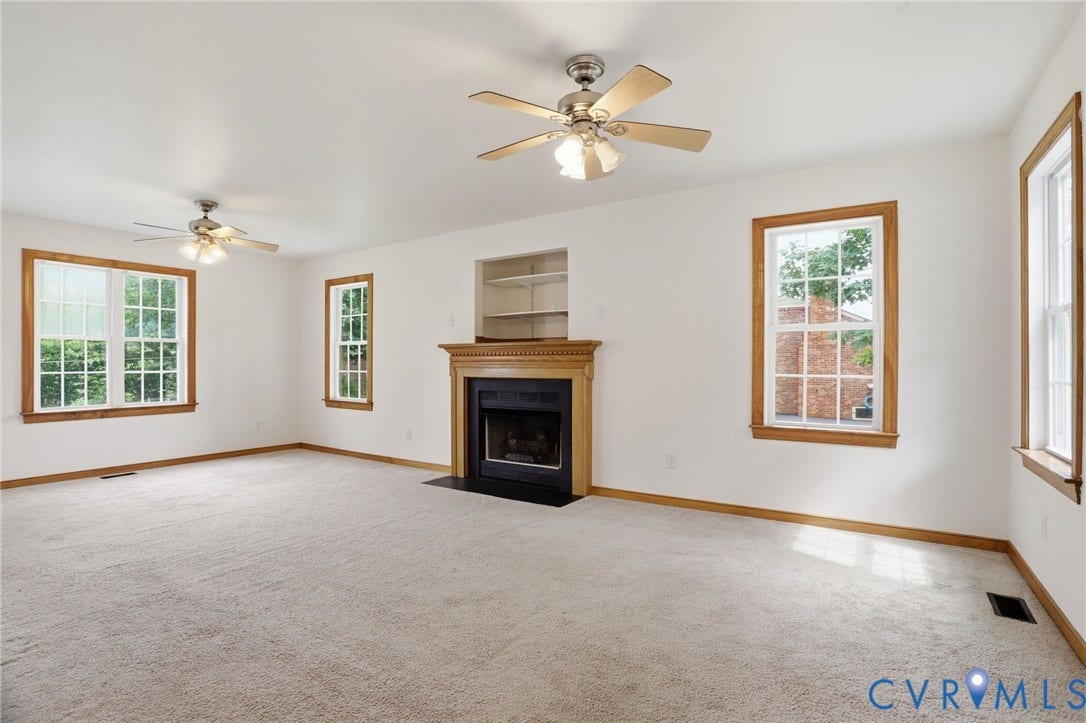 Spacious room featuring light-toned carpeting, multiple windows with wood-finish trim, a wood-mantled fireplace, and dual ceiling fans