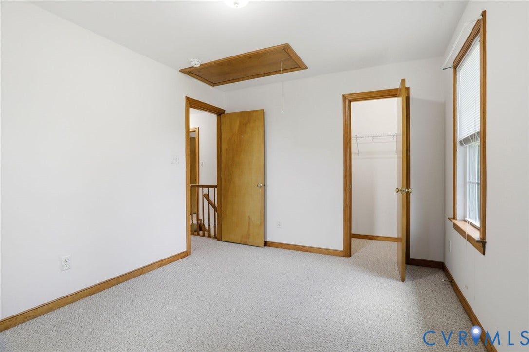 Carpeted room with white walls, featuring a window with blinds, wood-trim doorways, and a ceiling-mounted attic access panel