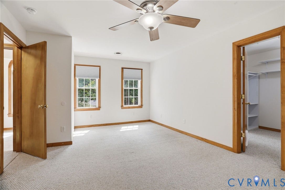 Spacious room featuring neutral carpeting, two windows with wood trim, a ceiling fan with integrated lighting, and a closet with shelving
