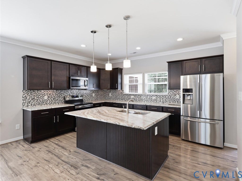 17431 Memorial Tournament Drive, Wynstone Park, Moseley, VA 23120 The chef-inspired kitchen showcases rich espresso cabinetry, granite countertops, a stylish tile backsplash, stainless steel appliances, luxury vinyl plank flooring, and an oversized island with bar seating, all beautifully illuminated by pendant and recessed lighting.