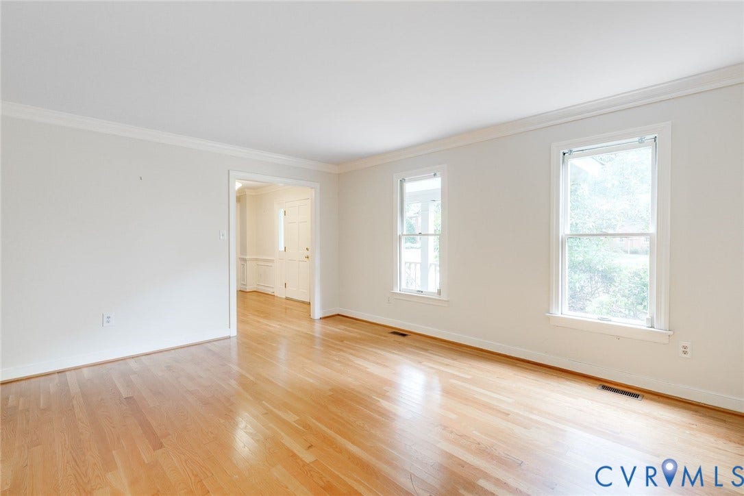 Hardwood flooring extends throughout this expansive room, featuring two double-hung windows with white trim, crisp white walls, and crown molding