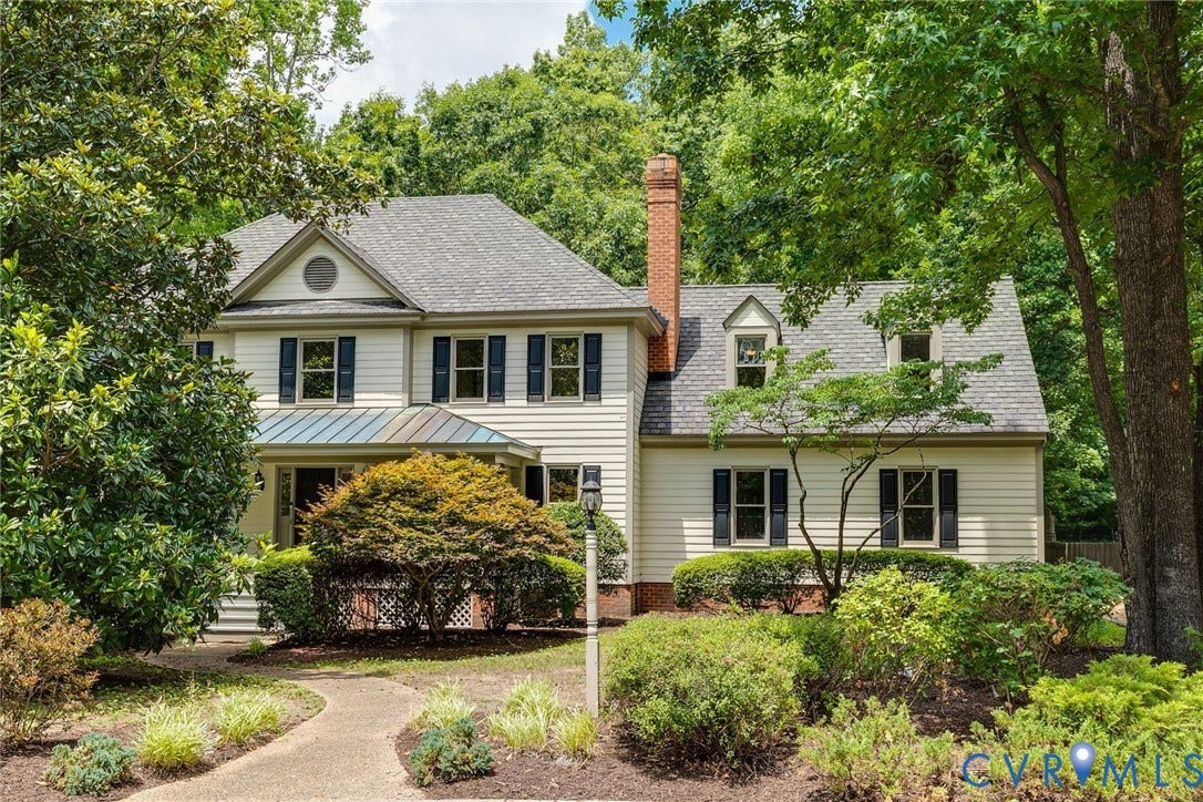 Traditional-style residence with light-colored siding, dark shutters, a prominent brick chimney, dormer windows, and a metal roof accent over the front entrance