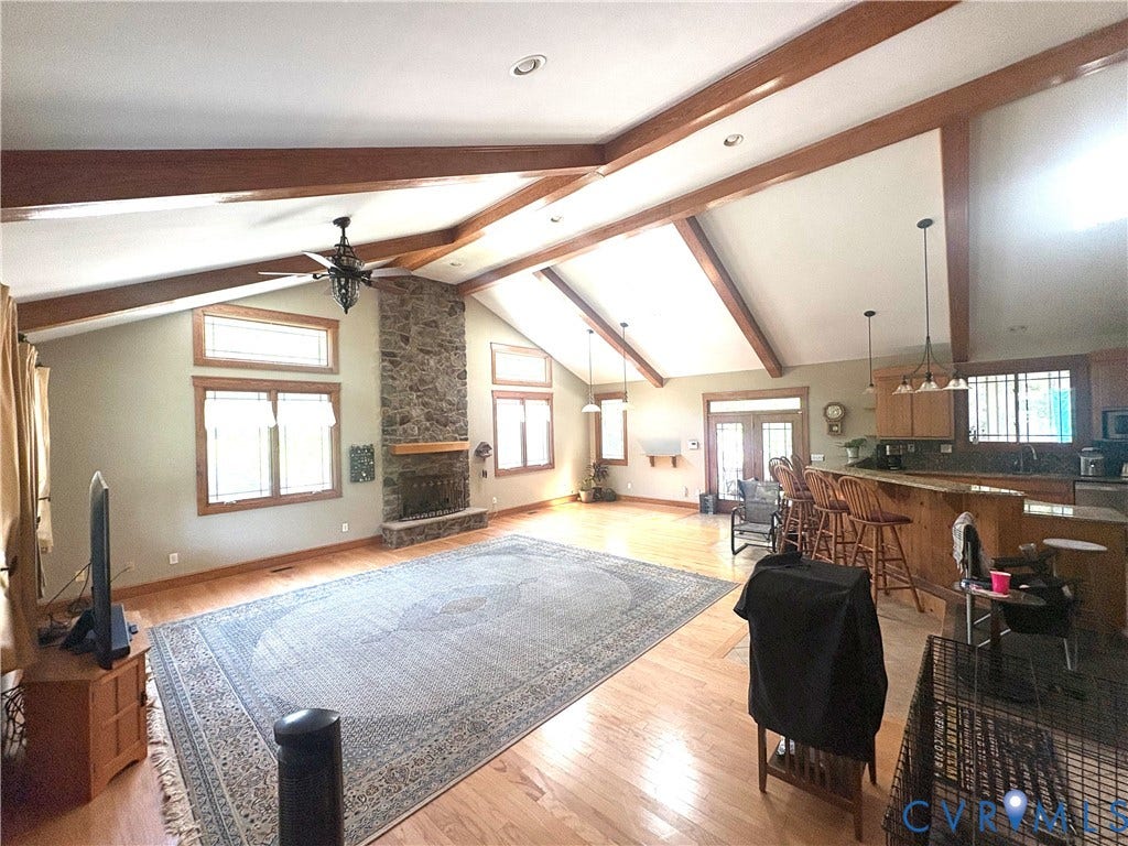 Expansive open-concept living area featuring vaulted ceilings with exposed wood beams, a stone-clad fireplace, and light wood-finish flooring