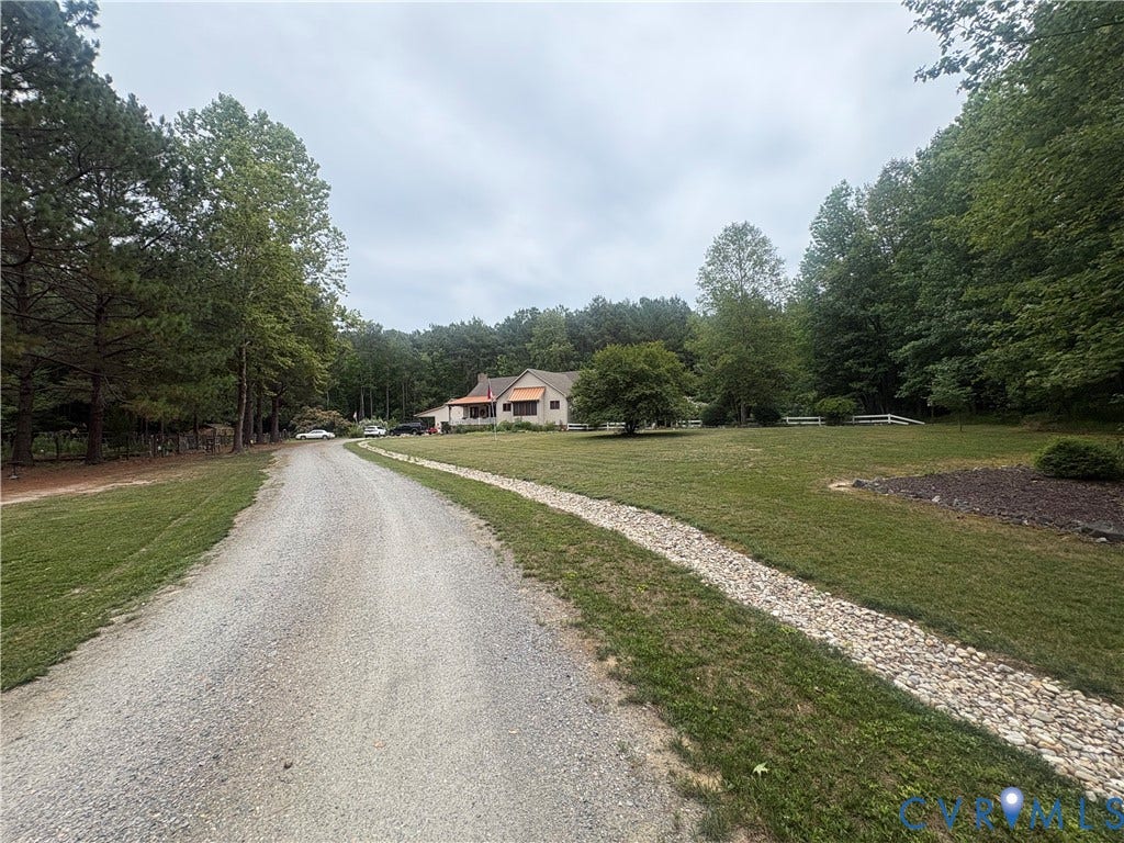 Gravel driveway leading to a ranch-style residence with a contrasting roof, surrounded by mature trees and a manicured lawn