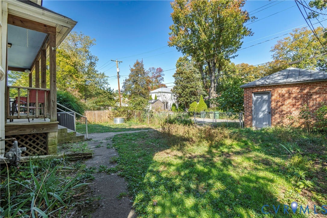 4404 Patterson Avenue, Richmond, VA 23221 Expansive outdoor space featuring a covered porch with wood railings and lattice skirting, a concrete pathway, a brick outbuilding with a gray door and shingle roof, and established trees