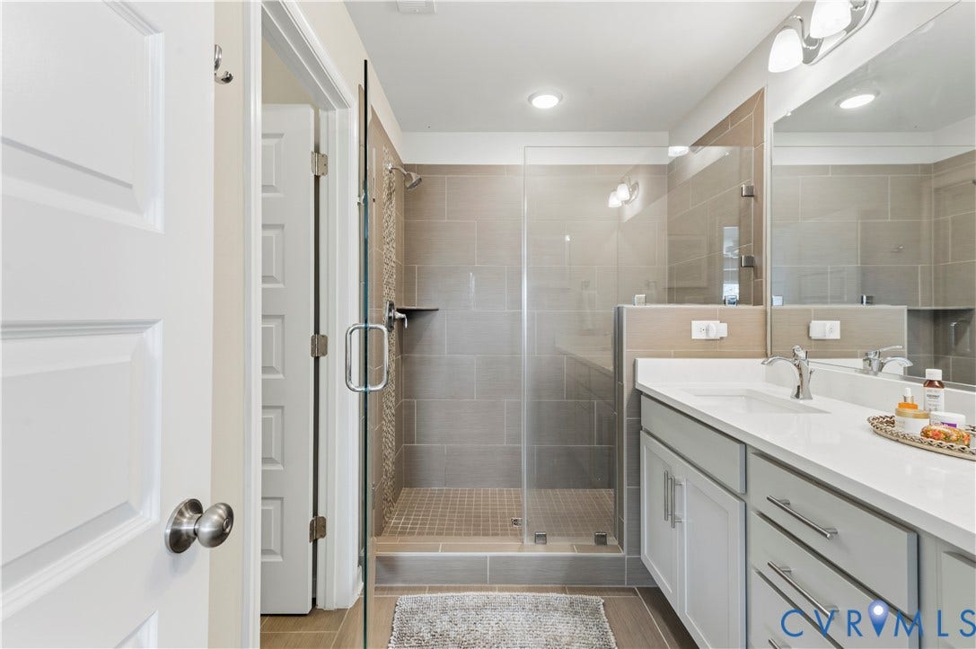 10414 Perennial Drive, Henrico, VA 23233 Spacious bathroom featuring a glass-enclosed shower with a tiled bench, a single vanity with a white countertop and undermount sink, light gray cabinetry with brushed nickel hardware, and wood-finish flooring