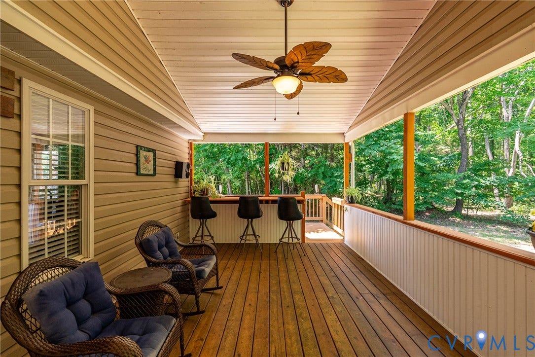 Spacious covered porch with wood-finish flooring, featuring a ceiling fan with leaf-shaped blades, a built-in bar with barstools, and a white beadboard railing