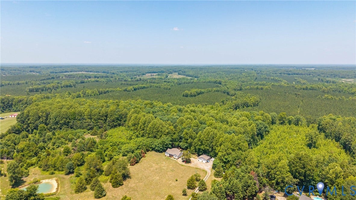 Expansive wooded landscape featuring a single-family residence, a detached auxiliary structure, a pond, and open fields