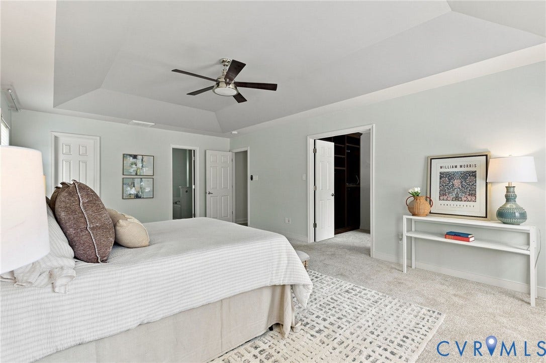 Tray ceiling with ceiling fan, light-colored wall paint, light-colored carpeting, and a doorway leading to a walk-in closet