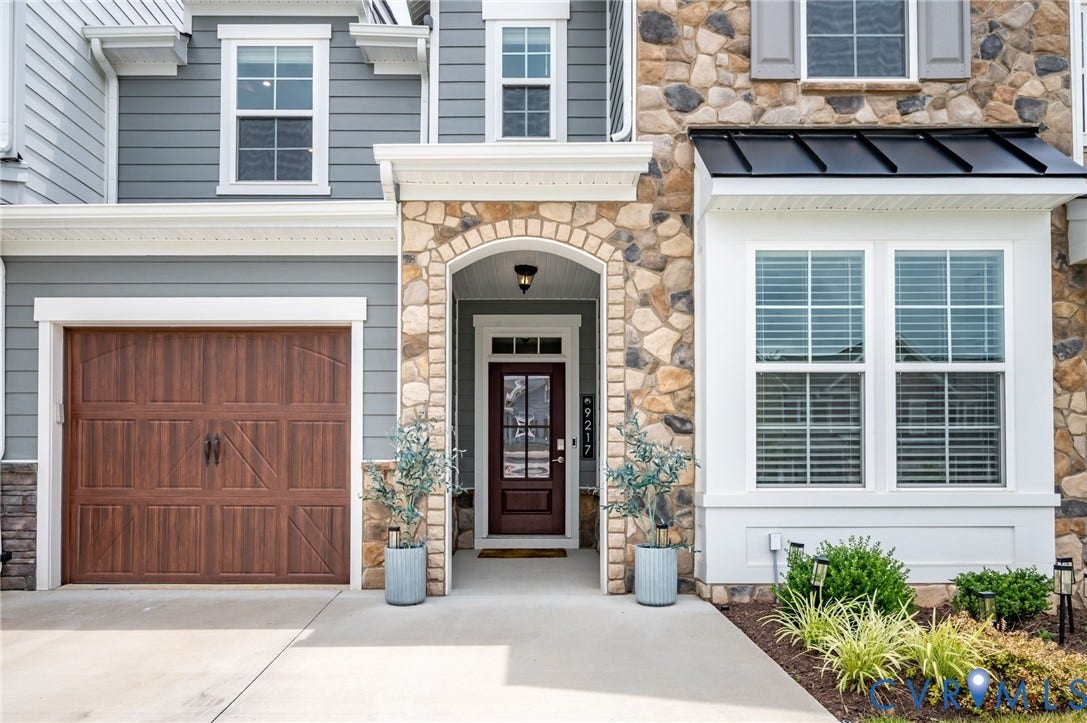 9217 Cerulean Place, Goochland, VA 23238 Exterior featuring stone and siding finishes, an arched entry with a glass-paneled door, a wood-finish garage door, and a bay window with a black metal awning
