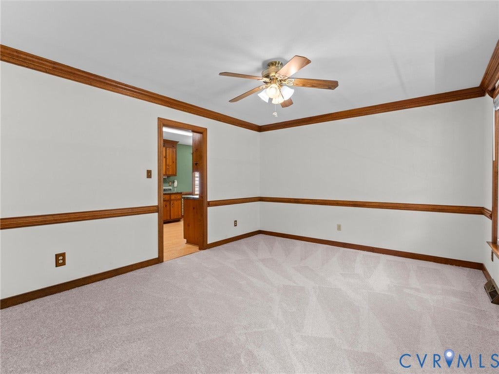 Carpeted room featuring white walls with wood trim and chair rail molding, a ceiling fan with integrated lighting, and a doorway leading to a kitchen with wood-finish flooring